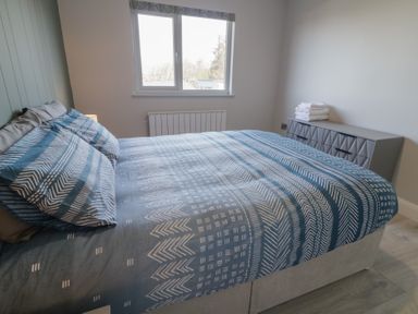 A bedroom with a bed with patterned blue bedding a window and a dresser with folded towels at Number 2 Villa - The View in Derrybeg County Donegal