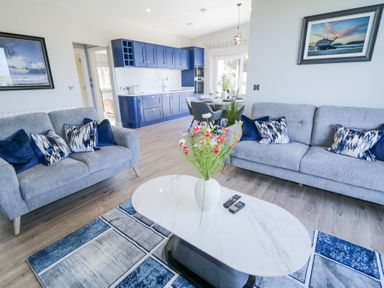 A living room with two grey sofas a white oval coffee table with flowers a blue and grey rug and a kitchen with blue cabinets at Number 5 Villa - The View in Derrybeg County Donegal