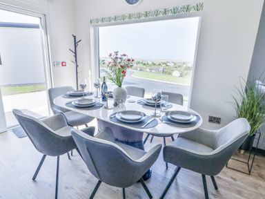 A dining area with a white oval table set for six with grey chairs and a vase of flowers by a large window at Number 5 Villa The View in Derrybeg County Donegal