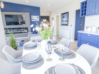 A dining table set with plates glasses and cutlery next to a kitchen with blue cabinets and a living room with a television and fireplace at Number 5 Villa - The View in Derrybeg County Donegal
