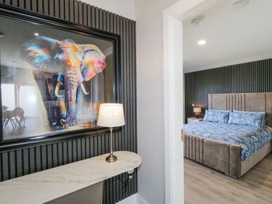A bedroom with a large bed and bedside tables seen through a doorway and a hallway with a colorful elephant painting and a lamp on a console table at Number 5 Villa - The View in Derrybeg County Donegal