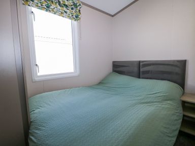 A bedroom with a bed covered by a green textured blanket a small bedside table and a window with green and yellow leaf patterned blinds at HW42 in Bognor Regis