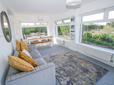 A living room with a gray sofa and yellow pillows a wooden dining table with chairs and large windows at Burry House in Llanmorlais