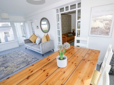 A dining area with a wooden table and white chairs next to a living room with a grey couch and yellow cushions at Burry House in Llanmorlais