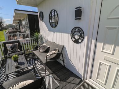 A small outdoor seating area with black chairs, a table with a plant, decorative wall hangings, and a white door at 56 Cherry Park Chalet in Chapel St Leonards