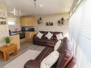 A living room with brown leather sofas next to a kitchen area with wooden cabinets and wall shelves at 56 Cherry Park Chalet in Chapel St Leonards