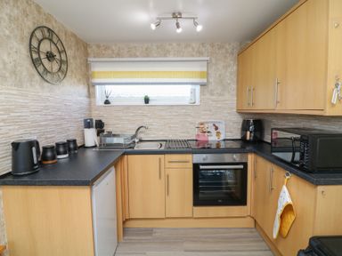 A kitchen with wooden cabinets a black countertop a sink oven microwave kettle and wall clock at 56 Cherry Park Chalet in Chapel St Leonards