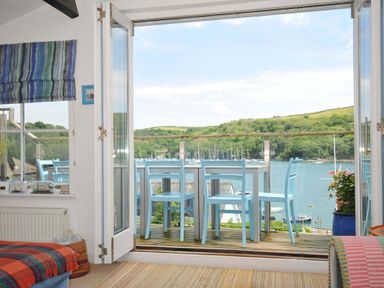 A room with an open glass door leading to a balcony with blue chairs and a table overlooking a river and green hills at 5 Esplanade in Fowey