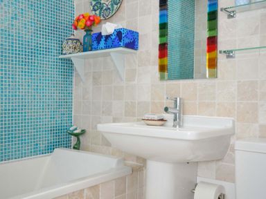A bathroom with a white bathtub and sink blue tiled wall a colorful mirror and a shelf with flowers and a tissue box at 5 Esplanade in Fowey