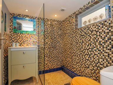 A bathroom with a sink, shower area with a glass partition, tiled walls, a toilet with a wooden seat, and a window with decorative shells at 5 Esplanade in Fowey