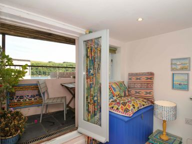 A small balcony with a chair and table next to a room with colorful cushions on a blue storage chest and a lamp on a small table at 5 Esplanade in Fowey