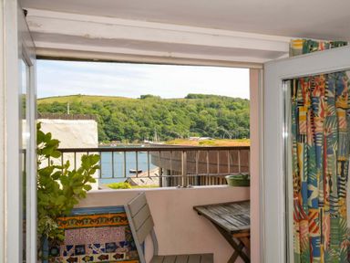 A balcony with a wooden table and chair overlooking a body of water and green hills at 5 Esplanade in Fowey