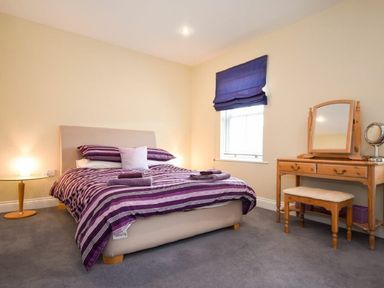A bedroom with a bed with striped bedding a round side table with a lamp and a wooden dressing table with a mirror and stool at Churchside in Alnwick