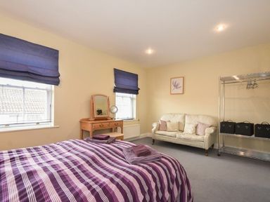 A bedroom with a bed covered in a purple striped blanket a wooden dressing table with a mirror two windows with blue blinds a white sofa and a metal clothes rack at Churchside in Alnwick