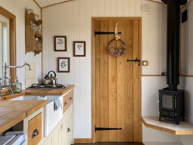 A kitchen with a wooden countertop a white sink a kettle on a stove a wooden door with a wreath and a black wood stove in the corner at Shepherd Hut 1 in Northchapel
