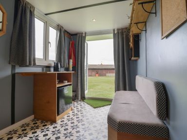 A small room with a bench seat a wooden cabinet with a kettle and fridge and curtains at Dozy Dotty Cow Shed 4 Easton near Campsea Ashe