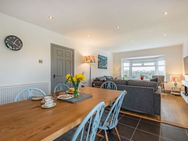A dining area with a wooden table and blue chairs next to a living room with grey sofas and a bay window at Bryn Derw in Caernarfon