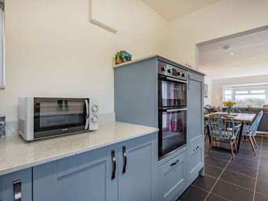A kitchen with blue cabinets a built-in double oven a microwave on the countertop and a dining area with chairs and a table with flowers at Bryn Derw in Caernarfon