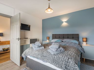 A bedroom with a bed towels on the blanket bedside tables with lamps a wall-mounted tv and an open door at Bryn Derw in Caernarfon