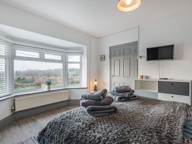 A bedroom with a bed covered in a gray textured blanket and folded towels a bay window with a vase and a cabinet with a wall mounted television at Bryn Derw in Caernarfon