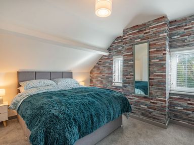 A bedroom with a double bed with teal fur blanket and two pillows beside two small windows with blinds and a wall mirror at Bryn Derw in Caernarfon