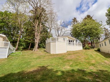 A mobile home with a white deck on a grassy hill surrounded by trees at 155 Woodland Hall Lyons in Ruthin