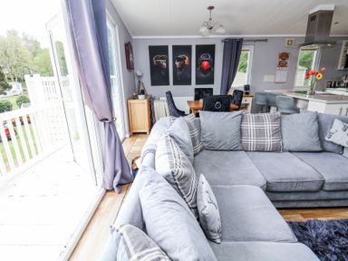 A living room with a large grey sectional sofa and a dining table with chairs next to a kitchen area with bar stools and a countertop at 155 Woodland Hall Lyons in Ruthin