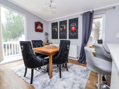 A dining area with a wooden table surrounded by black chairs on a gray rug beside a window with dark curtains at 155 Woodland Hall Lyons in Ruthin