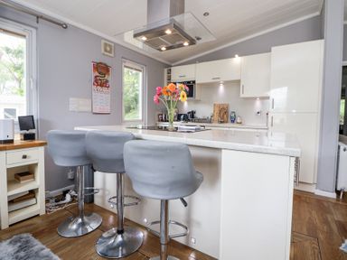 A kitchen with an island counter and three grey bar stools a vase with flowers on the counter white cabinets and wooden floor at 155 Woodland Hall Lyons in Ruthin
