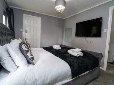 A bedroom with a bed with white linens and black throw blanket a wall-mounted television and a door at 155 Woodland Hall Lyons in Ruthin