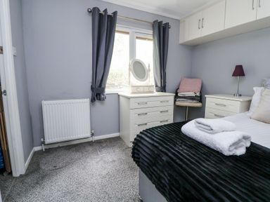A bedroom with a bed covered in a black blanket and white towels on it next to a white dresser with a mirror and a small table with a lamp at 155 Woodland Hall Lyons in Ruthin