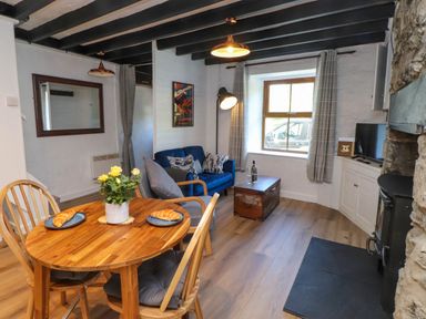 A living room with a wooden dining table and chairs croissants on plates and a vase with yellow flowers a blue sofa and an armchair a wooden chest a window with curtains a television and a fireplace at 4 Old Tanrhiw in Beddgelert