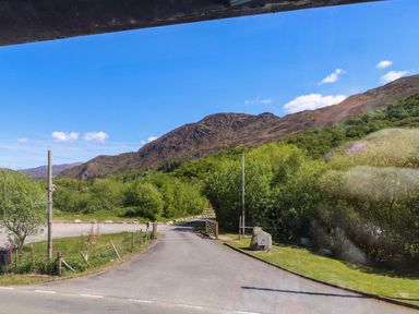 A paved road leading into a green area with trees and hills under a blue sky at 4 Old Tanrhiw in Beddgelert