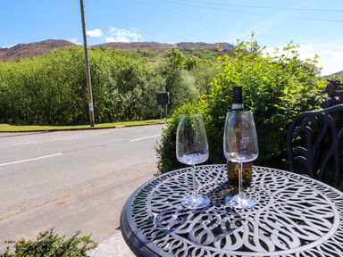 A metal outdoor table with two wine glasses and a bottle of wine next to a road with trees and hills in the background at 4 Old Tanrhiw in Beddgelert