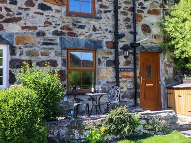 A stone house exterior with a wooden door a table and two chairs outside with wine glasses and greenery around at 4 Old Tanrhiw in Beddgelert