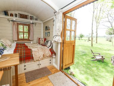 A small room with a built-in bed covered with blankets and pillows next to an open wooden door leading to a grassy yard with a wooden chair at Sika in Warmwell