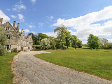 A gravel driveway leading to a stone building with multiple chimneys and windows surrounded by green lawn and trees at Sika in Warmwell