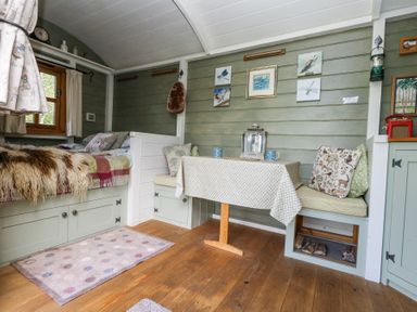A small room with a bed, a table with a tablecloth, bench seating with cushions, wall art, and a wooden floor at Fallow in Warmwell