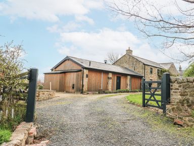 A gravel driveway leading to a stone and wooden barn with a dark gate and stone wall at The Barn in Riding Mill near Corbridge