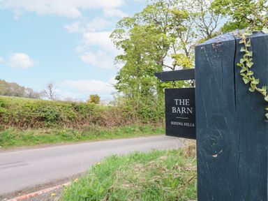 A roadside view with a wooden post and a sign reading The Barn Riding Hills surrounded by greenery