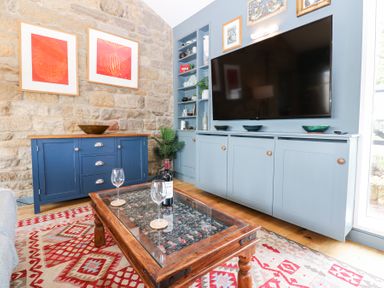 A living room with a TV on a blue cabinet a wooden coffee table with wine glasses and a bottle and a blue sideboard against a stone wall at The Barn in Riding Mill near Corbridge