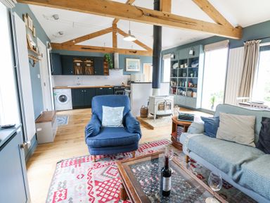 An open plan living room and kitchen with wooden beams a blue armchair a light blue sofa a wood stove and a kitchen with dark cabinets at The Barn Riding Mill near Corbridge