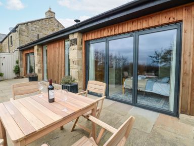 An outdoor patio with wooden table and chairs a wine bottle and glasses next to a stone and wood house with large glass doors at The Barn Riding Mill near Corbridge