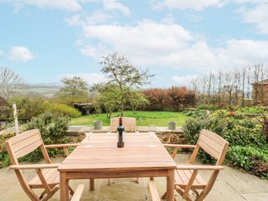 A wooden outdoor table with four chairs a bottle of wine and two glasses on a stone patio overlooking a garden at The Barn in Riding Mill near Corbridge