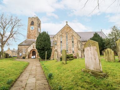 A stone church with a clock tower and a pathway leading to the entrance surrounded by grass and gravestones at The Barn Riding Mill near Corbridge