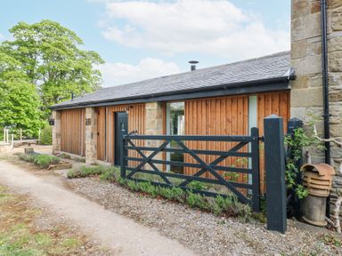 The exterior of a single story building with wooden paneling and a black gate along a gravel path at The Barn in Riding Mill near Corbridge