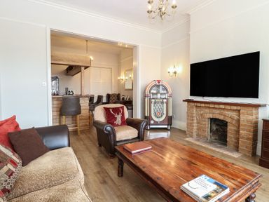 A living room with a sofa, armchair, wooden coffee table, jukebox, fireplace, and wall-mounted TV at The Burleigh in Hunstanton