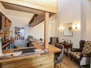 A small bar area with wooden counters stools and armchairs near a side table with framed pictures at The Burleigh in Hunstanton