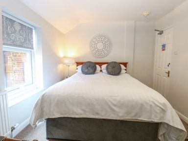 A bedroom with a large bed with white bedding and two round gray pillows a window with patterned blinds a table lamp and a decorative wall hanging above the bed at The Burleigh in Hunstanton