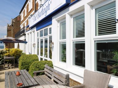 Outdoor seating area with wooden tables, benches, and an umbrella outside a building with white framed windows at The Burleigh in Hunstanton